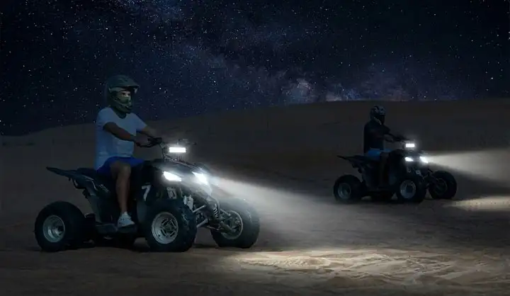 Two people riding quad bikes with bright headlights illuminated on desert sand dunes under a starry night sky with the Milky Way visible.
