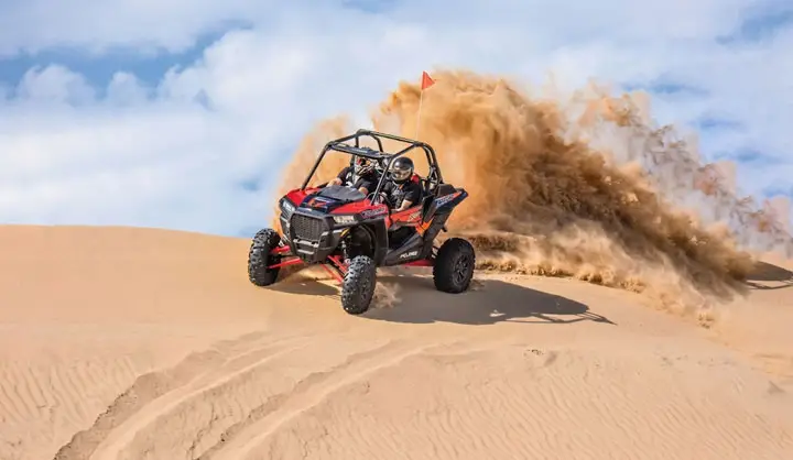 Red and black dune buggy speeding over a steep sand dune in Abu Dhabi desert, kicking up a large cloud of sand with passengers on board.
