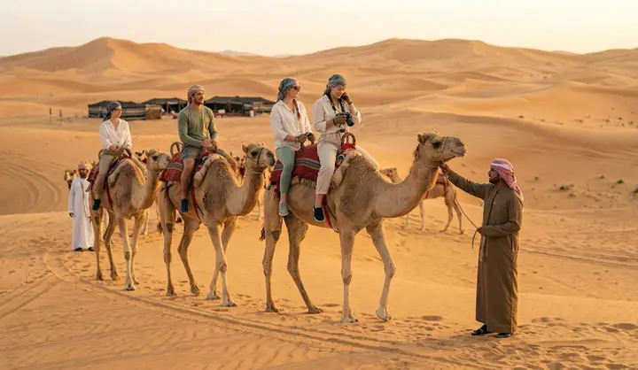 Tourists riding camels in a line through the golden sand dunes of Abu Dhabi desert, guided by a local camel handler at sunset.