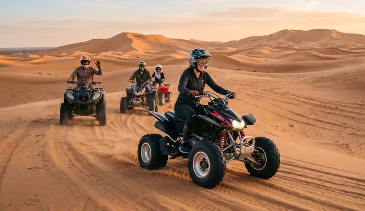 Group of adventurers riding quad bikes (ATVs) across golden sand dunes in the Abu Dhabi desert during a thrilling desert safari at sunset, with a woman in a black helmet leading the convoy.