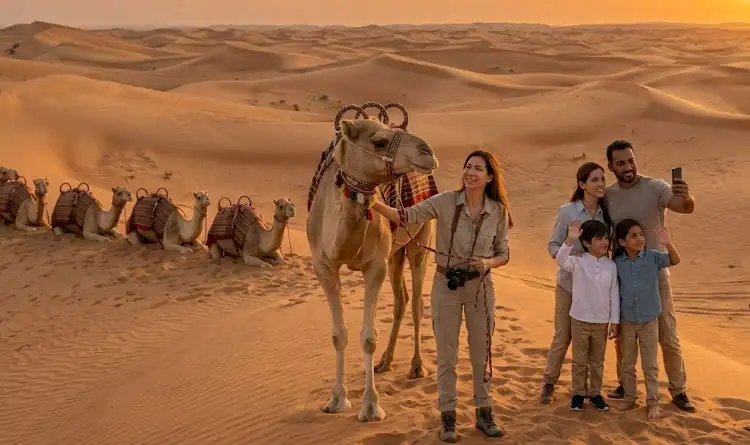 A smiling family of four takes a selfie with their smartphone next to a standing camel in the desert dunes during a warm sunset.