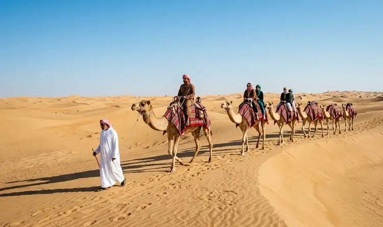 A guide in a white kandura leads a long line of tourists riding camels across rolling desert dunes under a bright, clear blue sky.