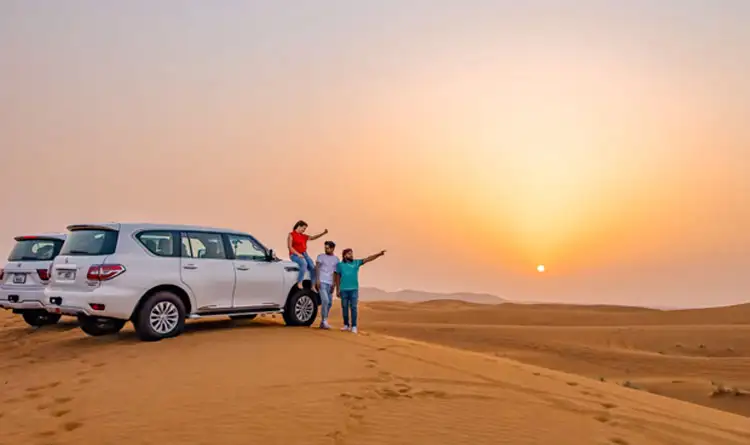 Friends standing on white SUV roof in desert at sunset, pointing at the view, golden dunes and dramatic sky.