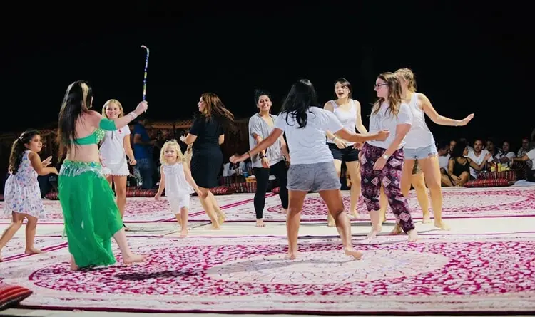 Guests dancing with a belly dancer on a stage carpet inside an evening desert camp in Abu Dhabi.