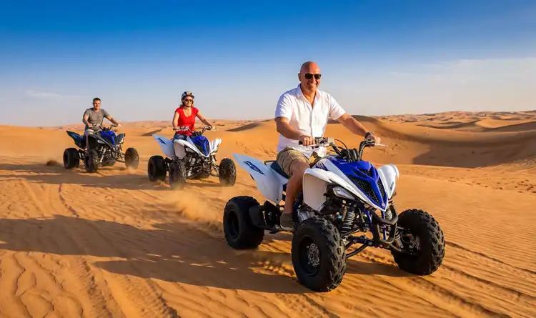 3 travellers riding exciting quad bikes in the controlled area of the Abu Dhabi desert, with a dune buggy in the background.