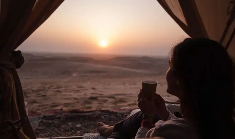Woman enjoying a peaceful sunrise with a cup of coffee inside a luxury desert tent, overlooking golden sand dunes in Abu Dhabi.