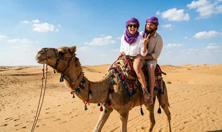 Smiling couple riding a decorated camel through the golden sand dunes of Abu Dhabi desert on a sunny day, both wearing purple headscarves and making peace signs.