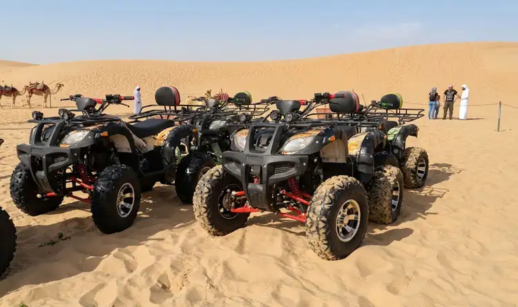 Four quad bikes parked on golden sand dunes in Abu Dhabi desert safari, with camels and people in the background under a clear blue sky.