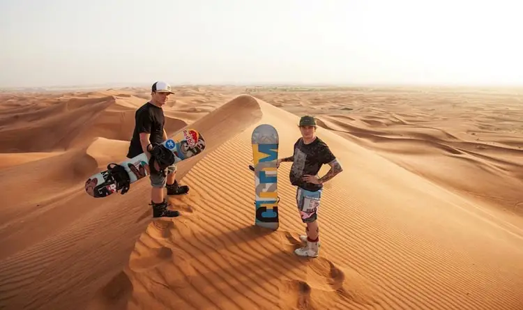 Two men standing with sandboards at the crest of a high sand dune during an Abu Dhabi desert safari.