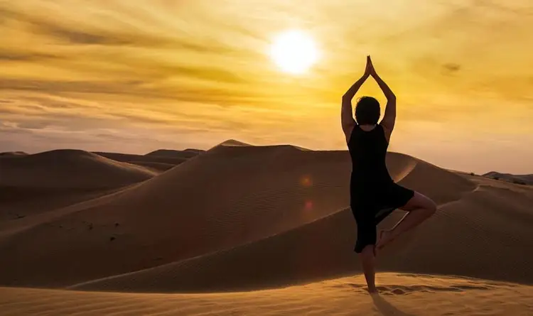 Woman practicing a tree pose yoga stance on top of sand dunes at sunrise in the Abu Dhabi desert.