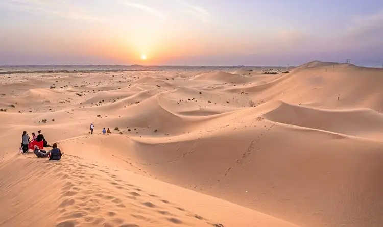 Group of tourists sitting on high sand dunes watching the sunrise over the vast Abu Dhabi desert landscape.